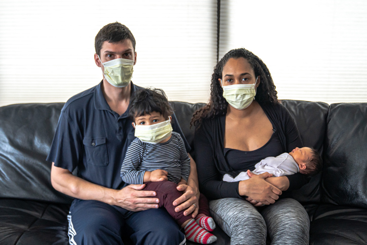 A mixed family sits on a black leather sofa with father and mother holding their infant son and daughter all wearing medical face masks in hopes of preventing getting sick from coronavirus